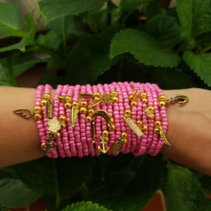 Pode incluir: Um conjunto de pulseiras de contas cor-de-rosa com pingentes dourados, incluindo uma pena, uma âncora, uma ferradura e um coração. As pulseiras são usadas no pulso.