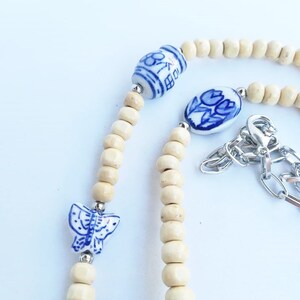 May include: A white and blue ceramic necklace with a butterfly, flower, and tulip design. The necklace is made with wooden beads and a silver chain.