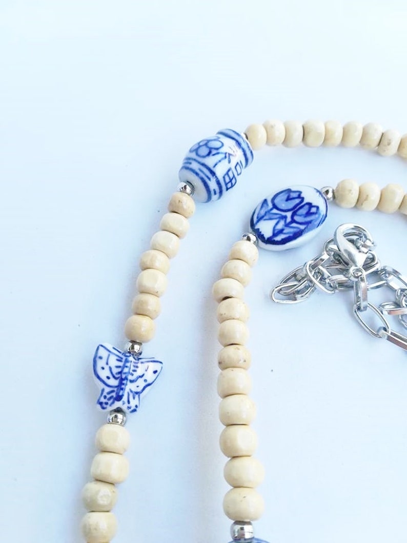 May include: A white and blue ceramic necklace with a butterfly, flower, and tulip design. The necklace is made with wooden beads and a silver chain.