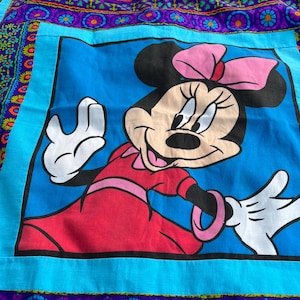Pu&ograve; includere: Un tessuto quadrato blu con un'immagine a cartoni animati di Minnie Mouse in un vestito rosso con un fiocco rosa. L'immagine &egrave; incorniciata da un bordo turchese e da un bordo viola e floreale a fantasia.