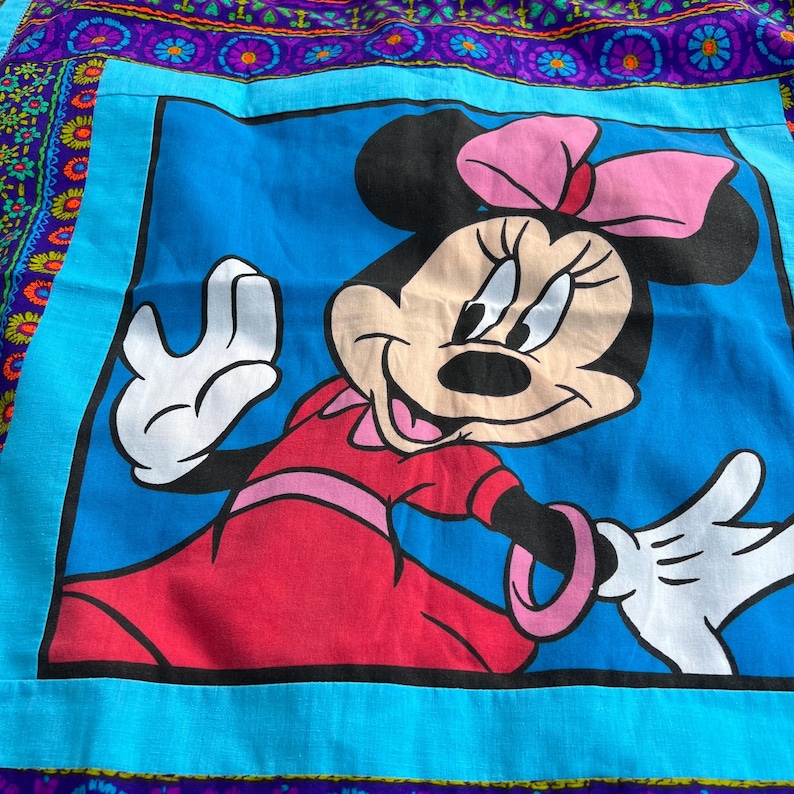 Pu&ograve; includere: Un tessuto quadrato blu con un'immagine a cartoni animati di Minnie Mouse in un vestito rosso con un fiocco rosa. L'immagine &egrave; incorniciata da un bordo turchese e da un bordo viola e floreale a fantasia.