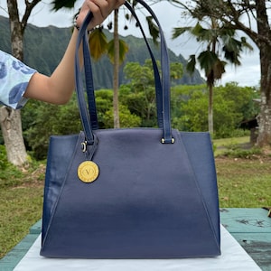 Op de afbeelding: Een marineblauwe leren tote bag met bijpassende handvatten en een goudkleurig medaillon. De tas heeft een rechthoekige vorm met een gestructureerd ontwerp. De handvatten zijn bevestigd met goudkleurige hardware.