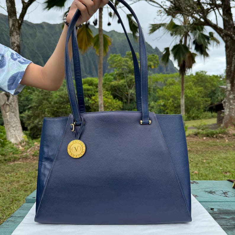 Op de afbeelding: Een marineblauwe leren tote bag met bijpassende handvatten en een goudkleurig medaillon. De tas heeft een rechthoekige vorm met een gestructureerd ontwerp. De handvatten zijn bevestigd met goudkleurige hardware.