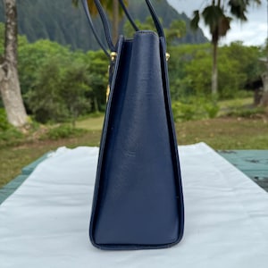 Op de afbeelding: Een marineblauwe handtas met een gestructureerd ontwerp. De tas heeft een rechthoekige vorm met een trapeziumvormig profiel. De handvatten zijn dun en donker, en de hardware lijkt goudkleurig te zijn. De tas staat op een wit oppervlak.
