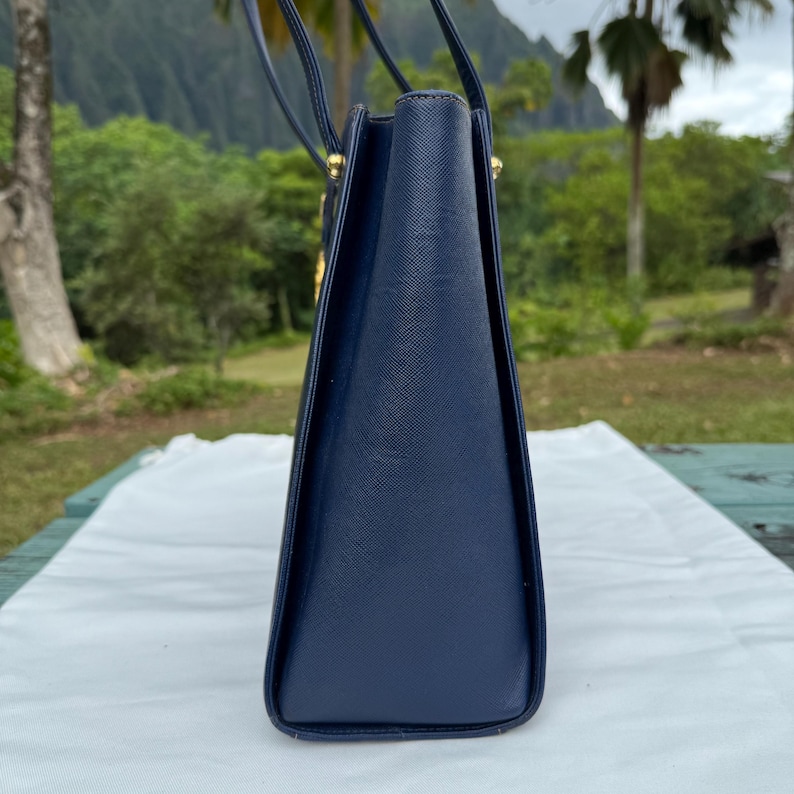 Op de afbeelding: Een marineblauwe handtas met een gestructureerd ontwerp. De tas heeft een rechthoekige vorm met een trapeziumvormig profiel. De handvatten zijn dun en donker, en de hardware lijkt goudkleurig te zijn. De tas staat op een wit oppervlak.