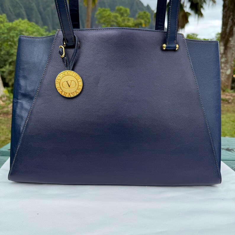 Op de afbeelding: Een marineblauwe tote bag met een goudkleurige cirkelvormige bedel. De tas heeft twee handvatten en een gestructureerd ontwerp. De bedel heeft de letter "V" in het midden. De tas is gemaakt van een textuurrijk materiaal.