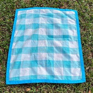 Pu&ograve; includere: Una coperta da picnic quadrata e trapuntata con un motivo a quadri bianchi e turchesi. La coperta ha un bordo turchese brillante ed &egrave; esposta su erba verde.