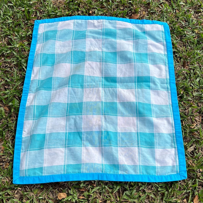 Pu&ograve; includere: Una coperta da picnic quadrata e trapuntata con un motivo a quadri bianchi e turchesi. La coperta ha un bordo turchese brillante ed &egrave; esposta su erba verde.