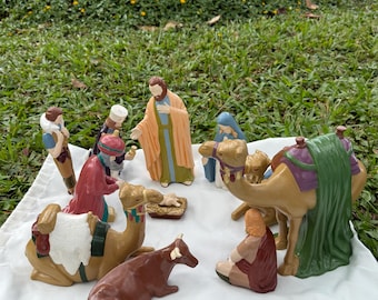 May include: A ceramic nativity scene with figures of Mary, Joseph, the baby Jesus, the three wise men, and animals. The figures are painted in various colors, including brown, red, blue, and gold. The scene is set on a white cloth.