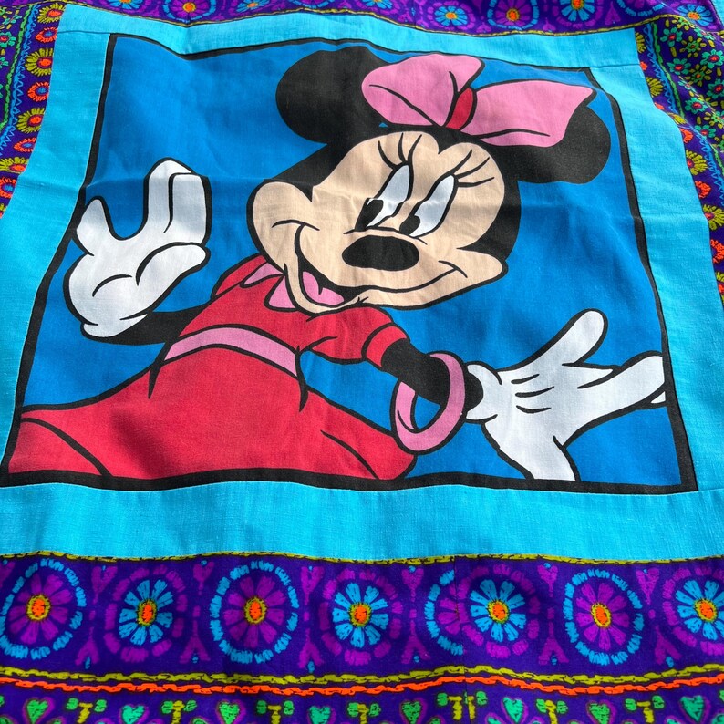 Pu&ograve; includere: Un tessuto quadrato con un'immagine a cartoni animati di Minnie Mouse con un vestito rosso e un fiocco rosa, su sfondo blu. Il bordo presenta un vivace mix di motivi floreali blu, viola e arancioni. Il design ha un'estetica vintage.
