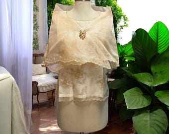 May include: A cream-coloured formal dress with a sheer shawl and a decorative brooch. The dress features floral embroidery and a scalloped lace trim. The outfit is displayed on a mannequin in front of a window with greenery.