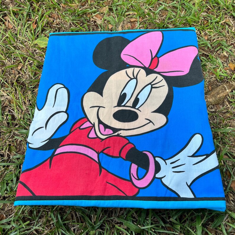 Pu&ograve; includere: Un telo mare blu con una grande grafica di Minnie Mouse. Minnie Mouse indossa un vestito rosso e un fiocco rosa e saluta con una mano guantata di bianco. L'asciugamano ha un bordo nero e blu.
