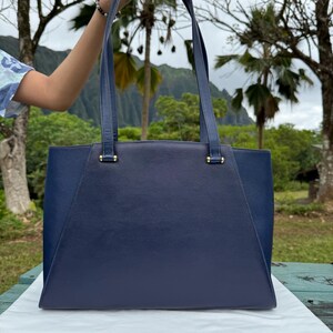 Op de afbeelding: Een marineblauwe draagtas met twee lange hengsels. De tas heeft een rechthoekige vorm met een gestructureerd ontwerp. De hengsels zijn met goudkleurige hardware aan de tas bevestigd. De achtergrond toont weelderige groene bomen en een bergketen.