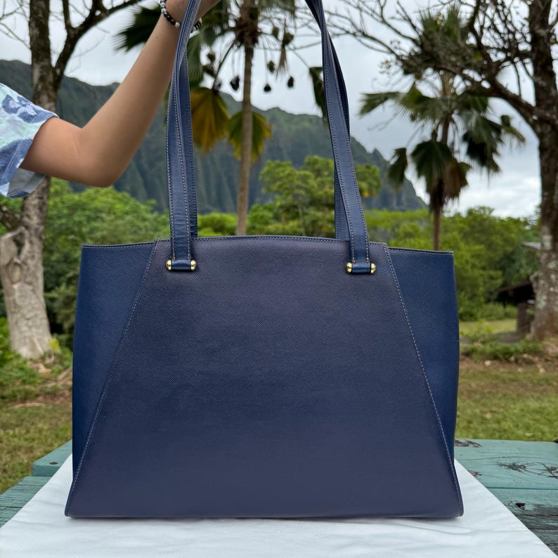 Op de afbeelding: Een marineblauwe draagtas met twee lange hengsels. De tas heeft een rechthoekige vorm met een gestructureerd ontwerp. De hengsels zijn met goudkleurige hardware aan de tas bevestigd. De achtergrond toont weelderige groene bomen en een bergketen.