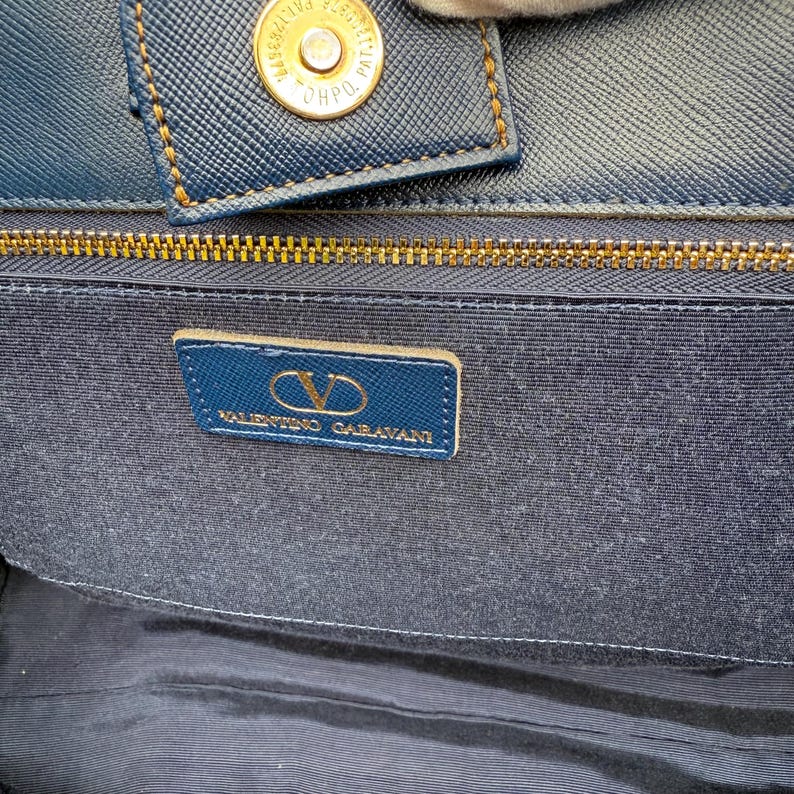 Op de afbeelding: Close-up van de binnenkant van een blauwe handtas met een gouden rits en een klein rechthoekig label. Het label toont het Valentino Garavani logo en tekst. Het materiaal van de tas heeft een textuur.