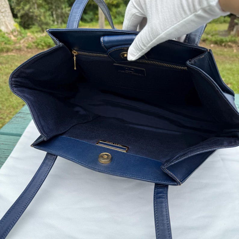 Op de afbeelding: Een marineblauwe tote bag met een gestructureerd ontwerp. De tas heeft twee handvatten, een goudkleurige sluiting en een binnenvak met ritssluiting. De binnenkant is gevoerd met bijpassende marineblauwe stof. De tas is open en onthult de binnenkant.