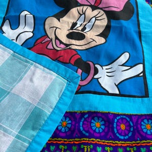 Pu&ograve; includere: Tessuto blu con un'illustrazione di Minnie Mouse in abito rosso e fiocco rosa. Il tessuto ha un bordo turchese e una sezione con un motivo floreale viola. &Egrave; visibile un angolo di tessuto a quadri.