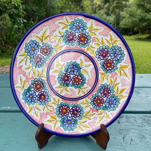 May include: A decorative ceramic plate with a blue rim and floral design. The plate features clusters of blue, purple, and maroon flowers with yellow leaves on a pink and white background. The plate is displayed on a wooden stand.