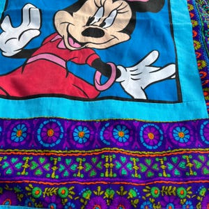Pu&ograve; includere: Tessuto vintage con una grande immagine di Minnie Mouse in un vestito rosso su sfondo blu. Il tessuto ha un bordo turchese e un motivo floreale decorativo viola e verde nella parte inferiore. Il design &egrave; colorato e stravagante.