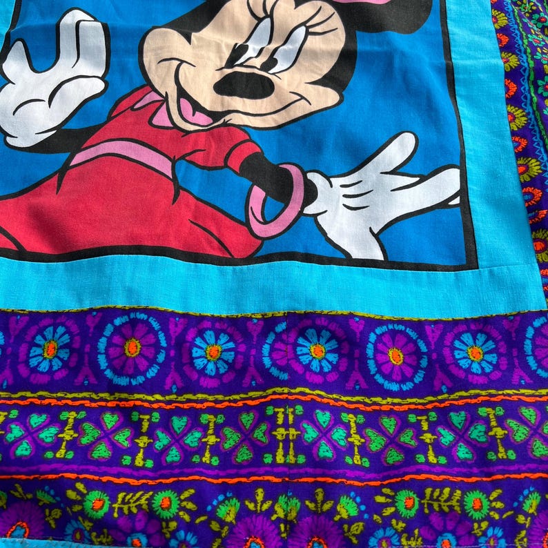 Pu&ograve; includere: Tessuto vintage con una grande immagine di Minnie Mouse in un vestito rosso su sfondo blu. Il tessuto ha un bordo turchese e un motivo floreale decorativo viola e verde nella parte inferiore. Il design &egrave; colorato e stravagante.