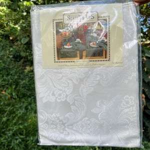 May include: A white jacquard table runner with a floral design, in its original packaging. The label reads "Statements" and shows a table setting. The runner is 178 cm long.