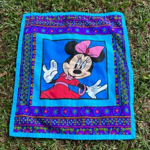Pu&ograve; includere: Un cuscino quadrato vintage con Minnie Mouse in abito rosso e fiocco rosa su sfondo blu. Il cuscino ha un bordo decorativo con motivi floreali e geometrici nei toni del viola, blu e verde.