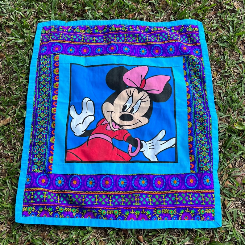Pu&ograve; includere: Un cuscino quadrato vintage con Minnie Mouse in abito rosso e fiocco rosa su sfondo blu. Il cuscino ha un bordo decorativo con motivi floreali e geometrici nei toni del viola, blu e verde.