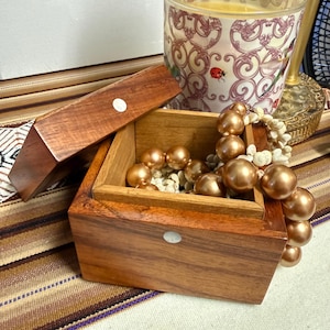 May include: A wooden jewelry box with a hinged lid, open to reveal gold-colored beads. A decorative candle in a glass jar with a floral design and a gold-colored beaded necklace are also present.