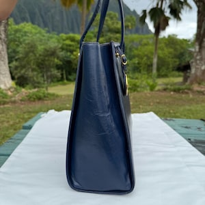 Op de afbeelding: Een marineblauwe leren tote bag met twee handvatten en goudkleurige hardware. De tas heeft een rechthoekige vorm en een glanzende afwerking. De tas staat op een wit oppervlak.