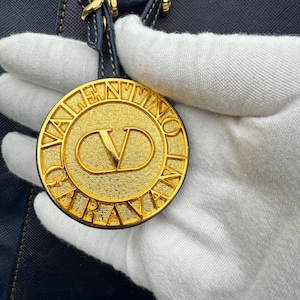 Op de afbeelding: Een goudkleurige, cirkelvormige bedel met de merknaam "VALENTINO" gegraveerd langs de rand. Het midden heeft een gestileerd "V"-logo. De bedel is bevestigd aan een marineblauwe leren band.