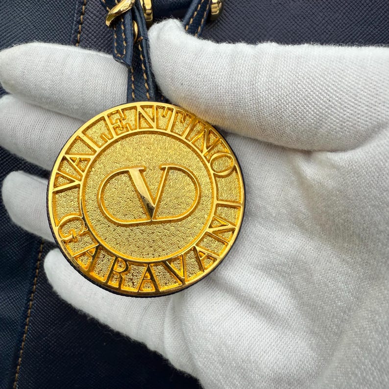 Op de afbeelding: Een goudkleurige, cirkelvormige bedel met de merknaam "VALENTINO" gegraveerd langs de rand. Het midden heeft een gestileerd "V"-logo. De bedel is bevestigd aan een marineblauwe leren band.