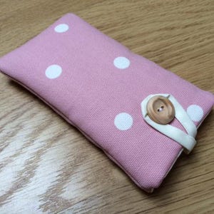 May include: A pink fabric pouch with white polka dots, secured by a wooden button and white strap. The rectangular case is likely designed to hold small items, such as keys or a phone.