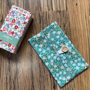 May include: A floral print tissue box and a matching fabric case with a button closure. The tissue box has a white background with red and pink roses and blue flowers. The fabric case is teal with a floral pattern.