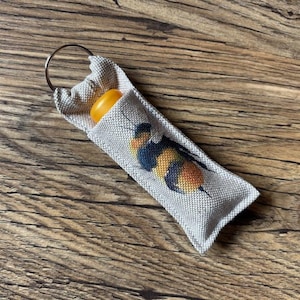 May include: A small, rectangular, light grey fabric pouch with a metal ring at the top. The pouch features a cross-stitched bumblebee design in yellow, black, and blue. An orange object is partially visible inside the pouch.