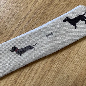 Peut inclure: Une trousse à crayons beige zippée avec un tirette de fermeture éclair blanche. La trousse présente deux silhouettes de teckel noires et deux silhouettes d'os blancs.