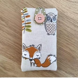 May include: A rectangular fabric phone case with a button closure. The case features a sleeping fox, an owl, and botanical illustrations in orange, brown, and green on a light beige background. The button is pink, and the loop is green with white polka dots.