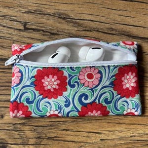May include: A small zipped pouch with a floral print in shades of red, pink, green, and blue. The pouch is open and contains two white wireless earbuds.