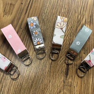 May include: Six fabric keychains with floral patterns and metal hardware are arranged on a wooden surface. The keychains feature various colors, including pink, blue, and white, with different floral designs. Each keychain has a metal ring.