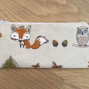 May include: A beige zippered pouch with a woodland theme. The pouch features a fox, an owl, mushrooms, acorns, butterflies, and pine trees.