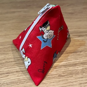 May include: A red triangular-shaped pouch with a zipper. The pouch is decorated with a repeating pattern of cartoon characters from the Peanuts comic strip, including Snoopy and Woodstock. The characters are depicted in various poses, including Snoopy flying his doghouse and Woodstock perched on his head. The pouch also features a blue star with a white outline. The text "Snoopy" is written in black cursive on the pouch.