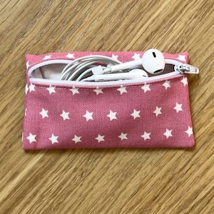 May include: A pink zippered pouch with white stars printed on it. The pouch is open and contains white earbuds.