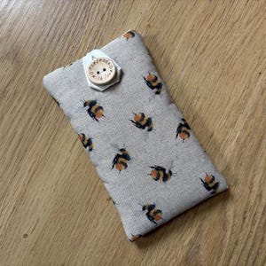 May include: A rectangular, light grey fabric case featuring a pattern of black and yellow bees. It is fastened with a wooden button that reads "handmade with love". The case is placed on a wooden surface.