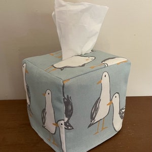 May include: A light blue tissue box cover with a white tissue box inside. The cover has a pattern of white seagulls with orange beaks on a light blue background.