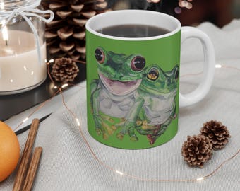 May include: White ceramic mug with a green background featuring a watercolour painting of two green frogs with red eyes. The mug is filled with a dark liquid and has a white handle.