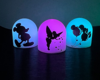 May include: Three night lights with different Disney character silhouettes. The first light is blue and features Mickey Mouse. The second light is purple and features Tinkerbell. The third light is blue and features Mickey Mouse with balloons.