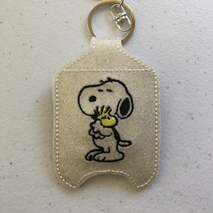 May include: A white glitter hand sanitizer holder with a black embroidered image of Snoopy holding a yellow bird. The holder has a silver metal key ring.