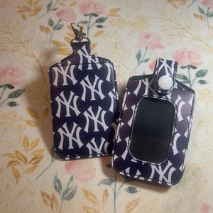 May include: Two navy blue and white patterned accessories, one with a silver clasp and the other with a white button closure. The pattern features the interlocking "NY" logo. The items are displayed on a floral background.