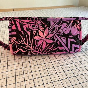 May include: A quilted fabric bag with a vibrant floral pattern in shades of pink and black. The bag features two curved handles and a rectangular body, perfect for carrying essentials. The design includes stylized flowers and leaves.