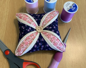 Puede incluir: Una bobina de hilo rosa, una bobina de hilo gris, una bobina de hilo morado, una aguja, unas tijeras y un alfiletero floral con un botón en el centro.