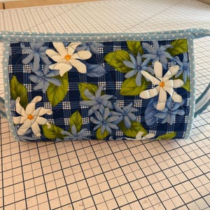 May include: A quilted fabric bag with a blue and white floral design. The bag features a navy blue plaid background with appliqued white and blue flowers and green leaves. It has light blue handles and trim.